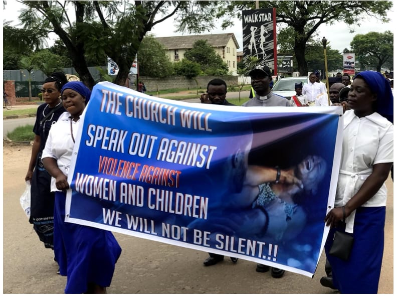 Two women in guild uniform carry a banner along a road reading “The church will speak out against violence against women and children. We will not be silent”. Clergy and other marchers follow behind.
