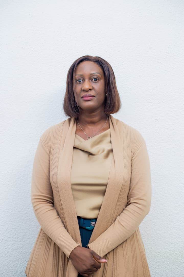 Womba M Lukwesa, in a tan cardigan over a matching top, photographed against a white wall.
