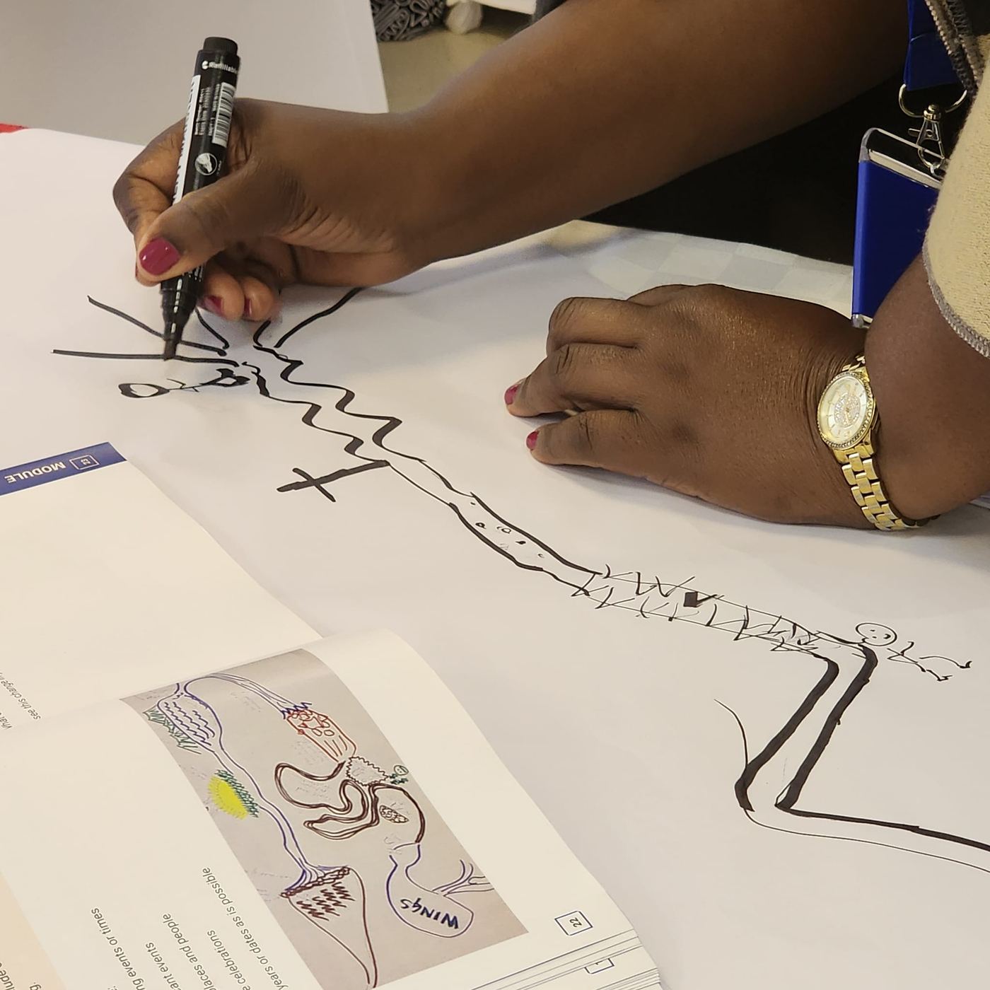A close view of hands drawing a root-cause diagram in marker on a sheet of flip-chart paper, beside a printed workbook page.